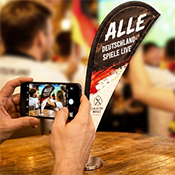 Hände einer Person, die mit ihrem Smartphone eine Tischfahne im WM-Design fotografiert, im Hintergrund sind jubelnde Menschen beim Public Viewing mit Deutschland-Fanartikeln zu sehen Hände einer Person, die mit ihrem Smartphone eine Tischfahne im WM-Design fotografiert, im Hintergrund sind jubelnde Menschen beim Public Viewing mit Deutschland-Fanartikeln zu sehen